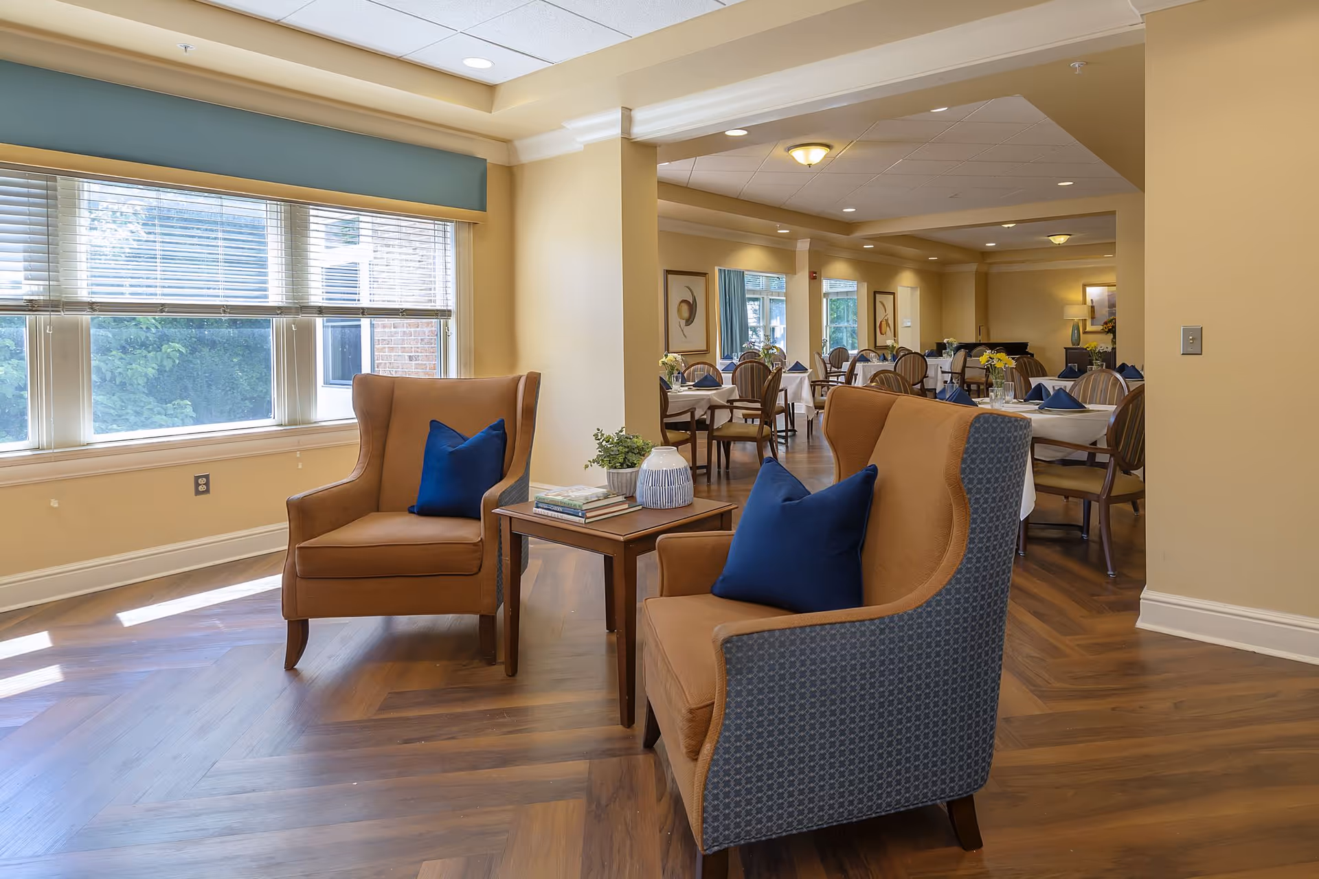 A cozy seating area with two brown armchairs featuring blue cushions and a small wooden table with books and a plant between them. Behind the seating area is a dining room with tables covered in white tablecloths and set with blue napkins. Large windows on the left let in natural light, and the room has warm beige walls and wooden flooring.
