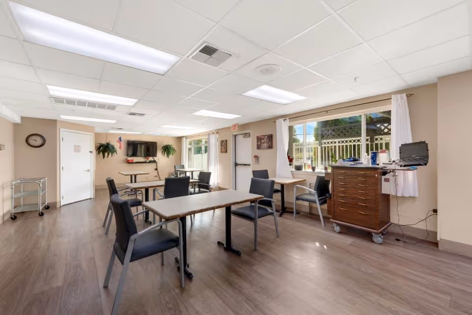 A bright and clean common room in a senior living facility with several tables and chairs arranged for group seating. The room has wood flooring, beige walls, and a large window with white curtains letting in natural light. There is a television mounted on the far wall, two potted plants hanging on either side of the TV, and a wooden cabinet with various items on top near the window. A walker is positioned near a closed door on the left side of the room.