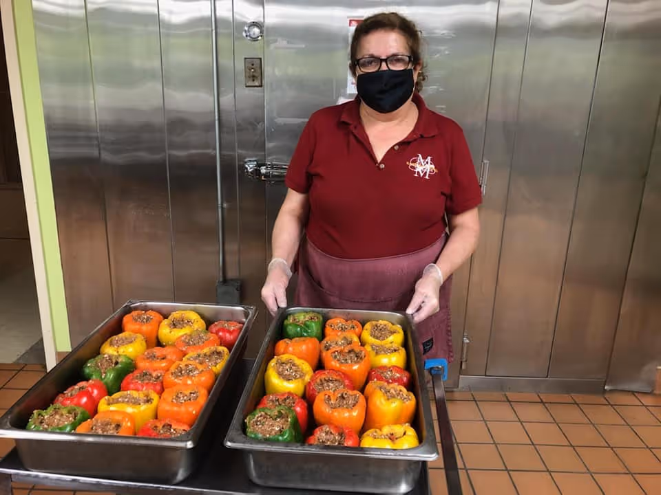 A masked staff member in a commercial kitchen holds two trays of colorful stuffed bell peppers.