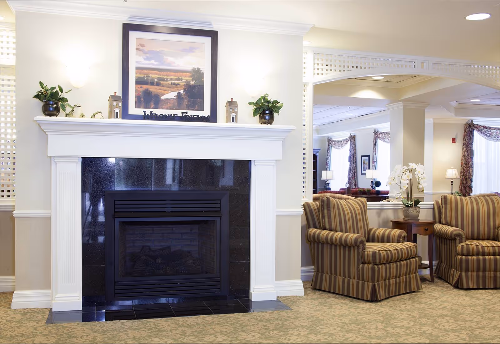 Cozy common room with a black-tiled fireplace, framed artwork above, and striped armchairs with a side table and orchid.