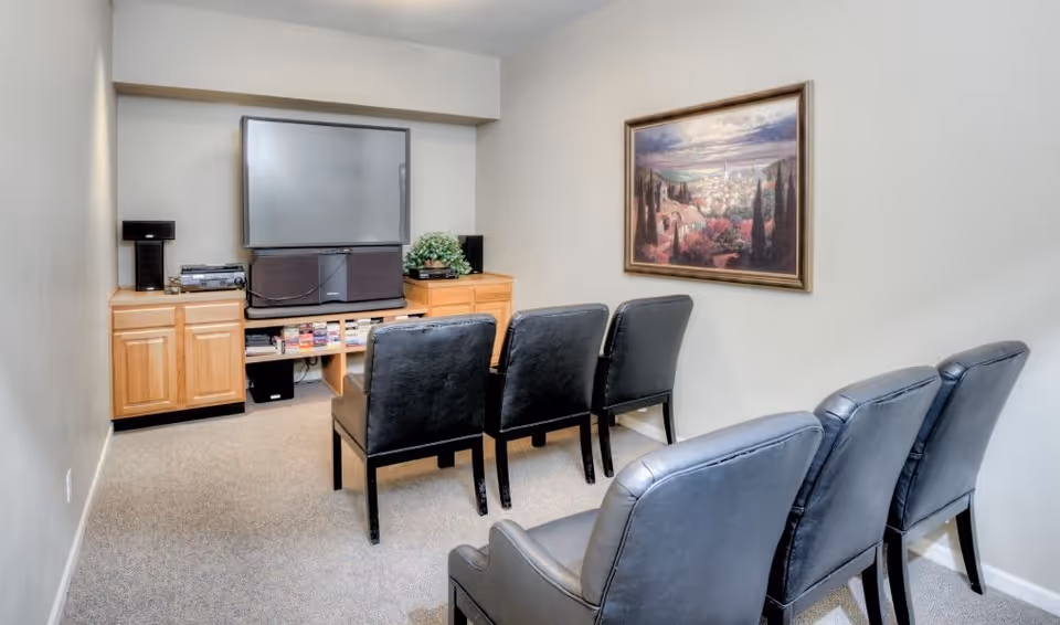 A small media room with five black leather chairs arranged in two rows facing a large flat-screen TV mounted on a wooden cabinet. The cabinet holds speakers, a stereo system, and some books. A framed landscape painting hangs on the wall to the right.