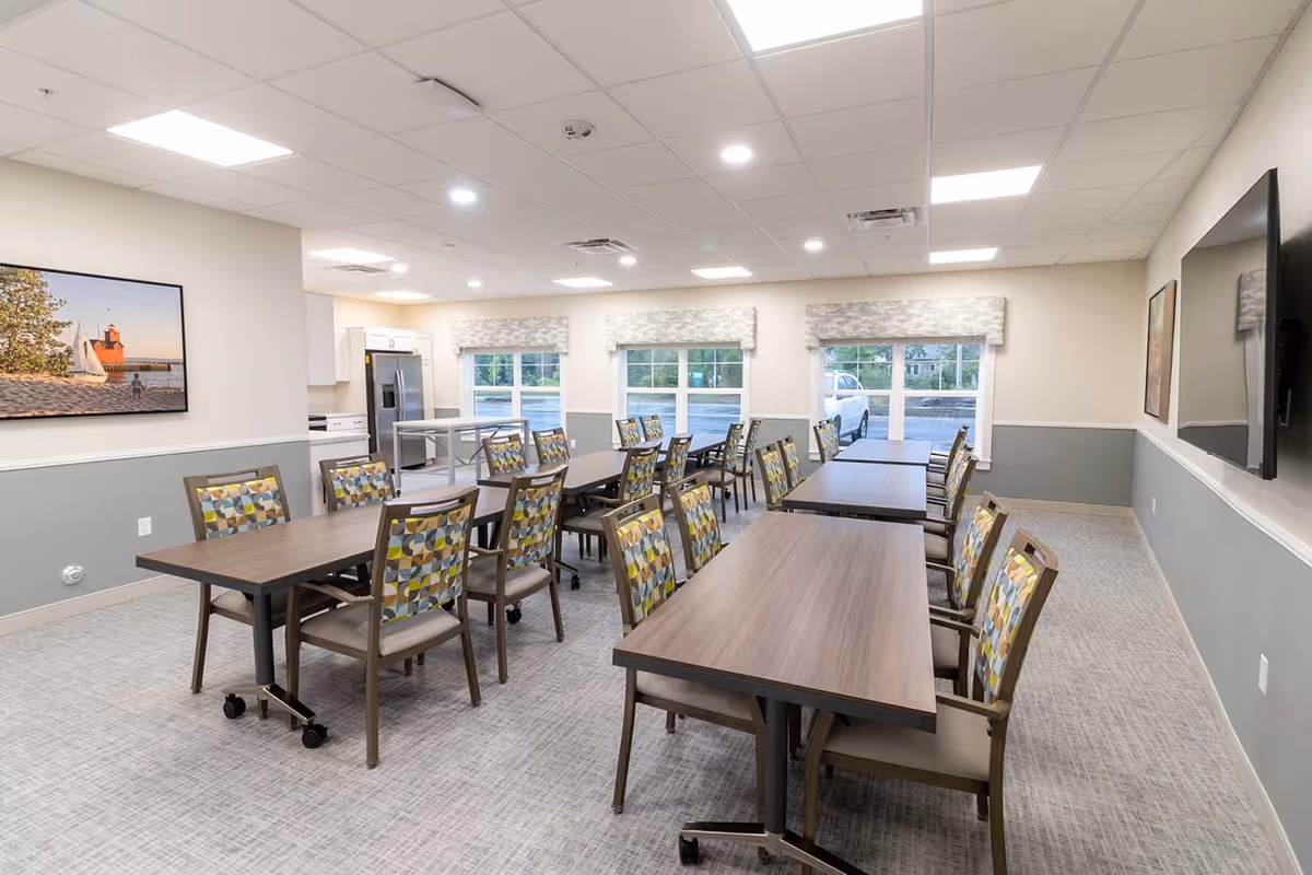 A bright and clean dining room in a senior living facility with several rectangular tables and chairs featuring patterned upholstery. The room has large windows with valances, a wall-mounted flat screen TV, and a kitchen area with a refrigerator and cabinets in the background. The walls are painted in light neutral colors with a chair rail and the floor is carpeted.