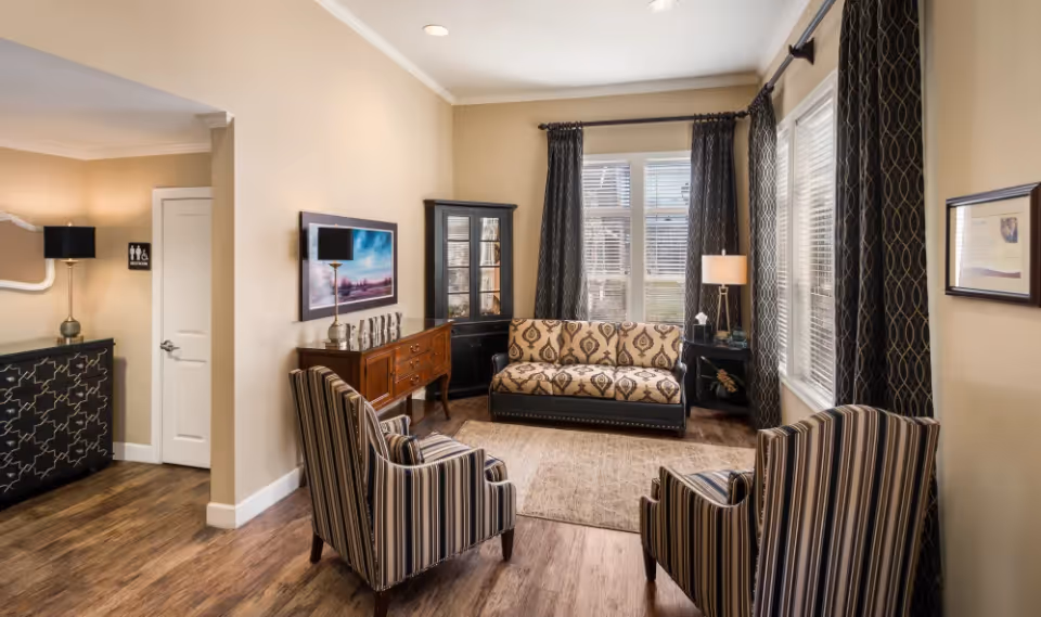 A cozy living room area with a patterned sofa, two striped armchairs, a wooden sideboard with decorative items, a black corner cabinet, and large windows with dark patterned curtains allowing natural light to fill the space. The room has beige walls, wooden flooring, and a floor lamp beside the sofa. A door and a black cabinet with a lamp are visible in the background.