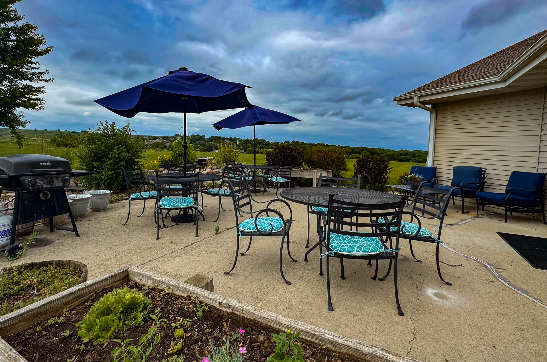 Outdoor patio with metal tables and chairs, blue umbrellas and cushions, a grill, and a view of green fields under a cloudy sky.