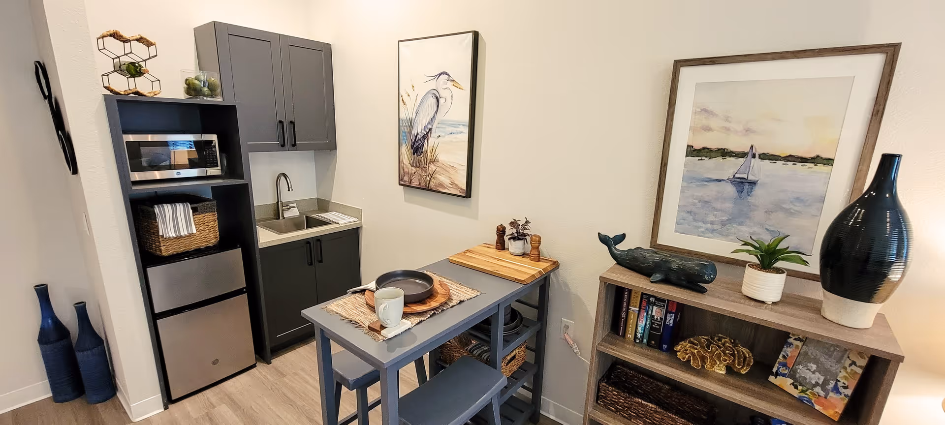 Small kitchenette and dining nook with a mini fridge, microwave, sink, narrow table and decorative shelving.