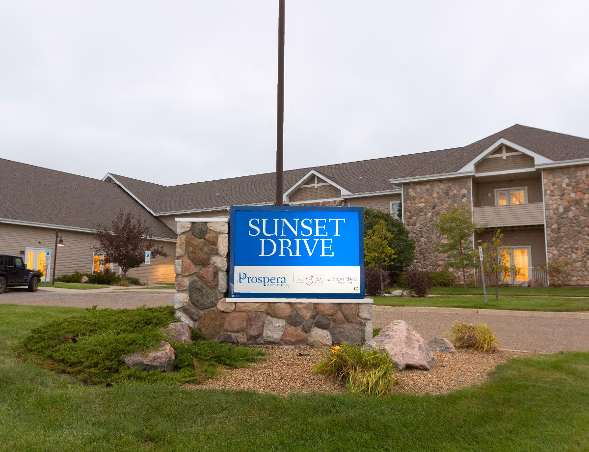 A stone monument sign reading 'SUNSET DRIVE' in front of a landscaped lawn and a two-story senior living building.