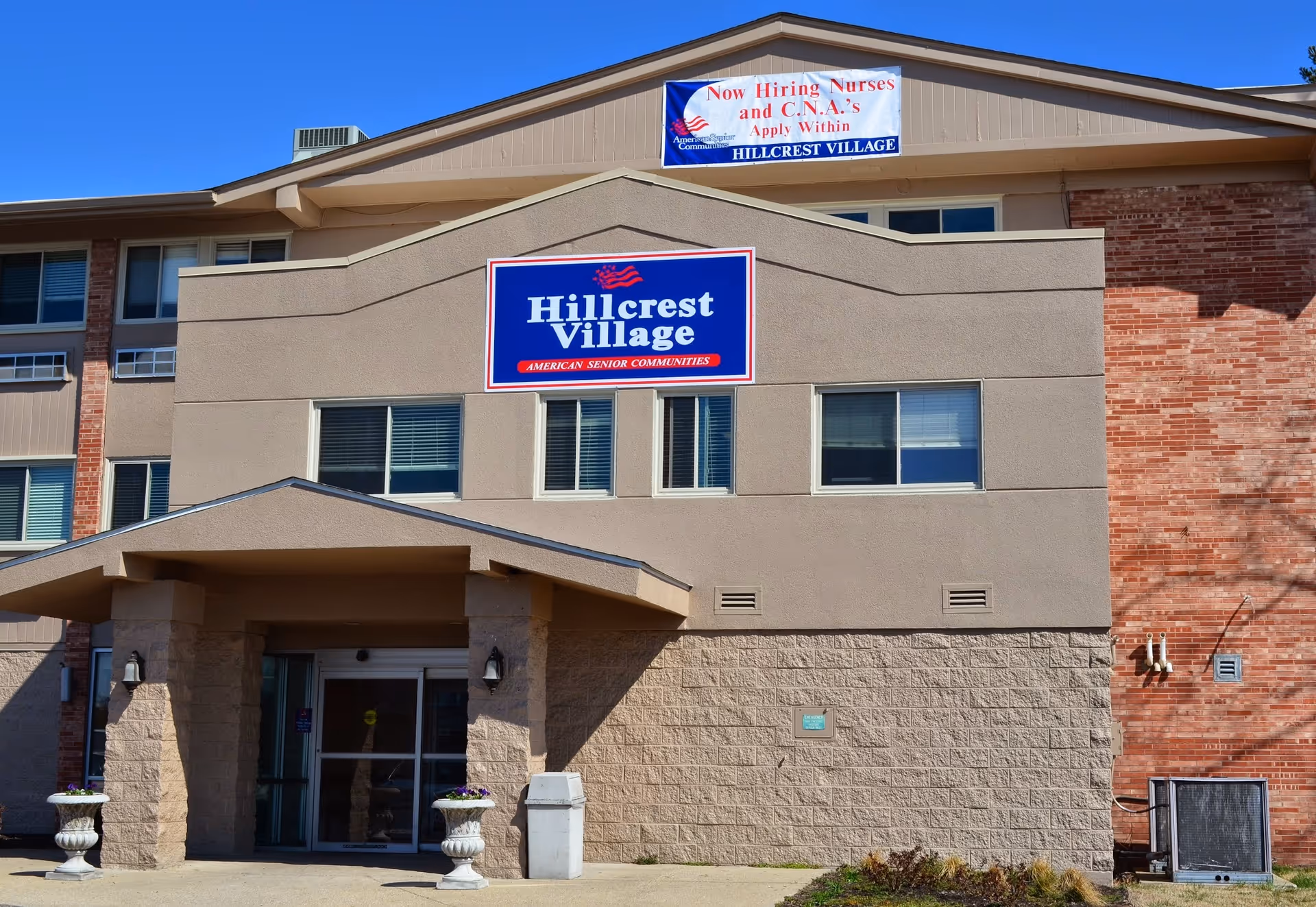 Exterior front view of Hillcrest Village senior living facility building with a beige and brick facade, a covered entrance, and signs displaying the facility name and a hiring notice for nurses and CNAs.