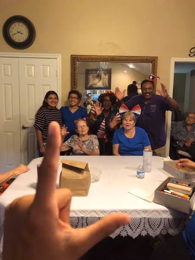 A group of people gathered around a table in a room, smiling and waving at the camera. The table is covered with a white lace tablecloth and has various items on it including a brown paper bag, a water bottle, and a box of colored pencils. A clock is visible on the wall above a closed door, and a large mirror reflects part of the room and a person taking the photo.