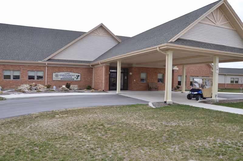 Front entrance of a senior living building with a covered porte-cochere, a sign reading 'Life Enrichment Center,' and a small parked golf cart.