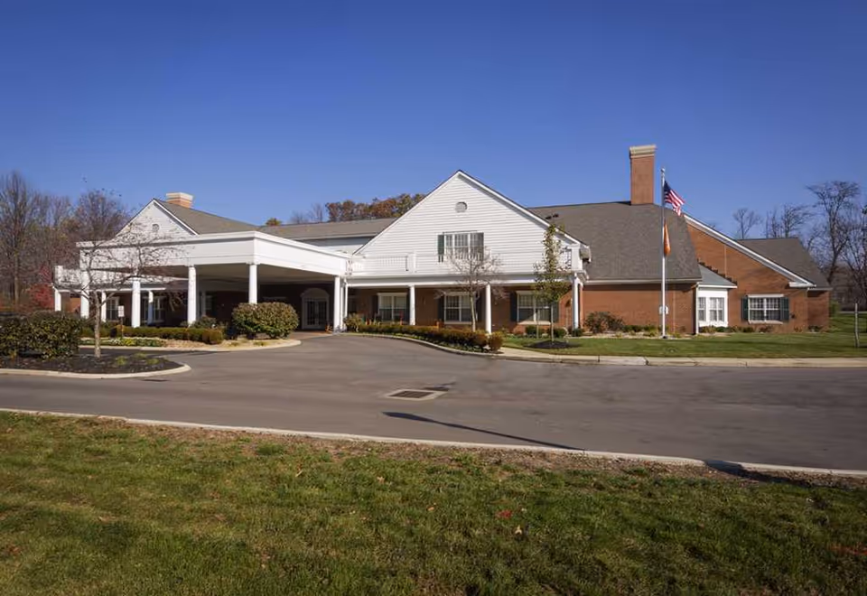 Exterior view of a single-story senior living facility building with a covered entrance, brick and white siding walls, and an American flag on a flagpole. The building is surrounded by a paved driveway, landscaped bushes, and a grassy area under a clear blue sky.