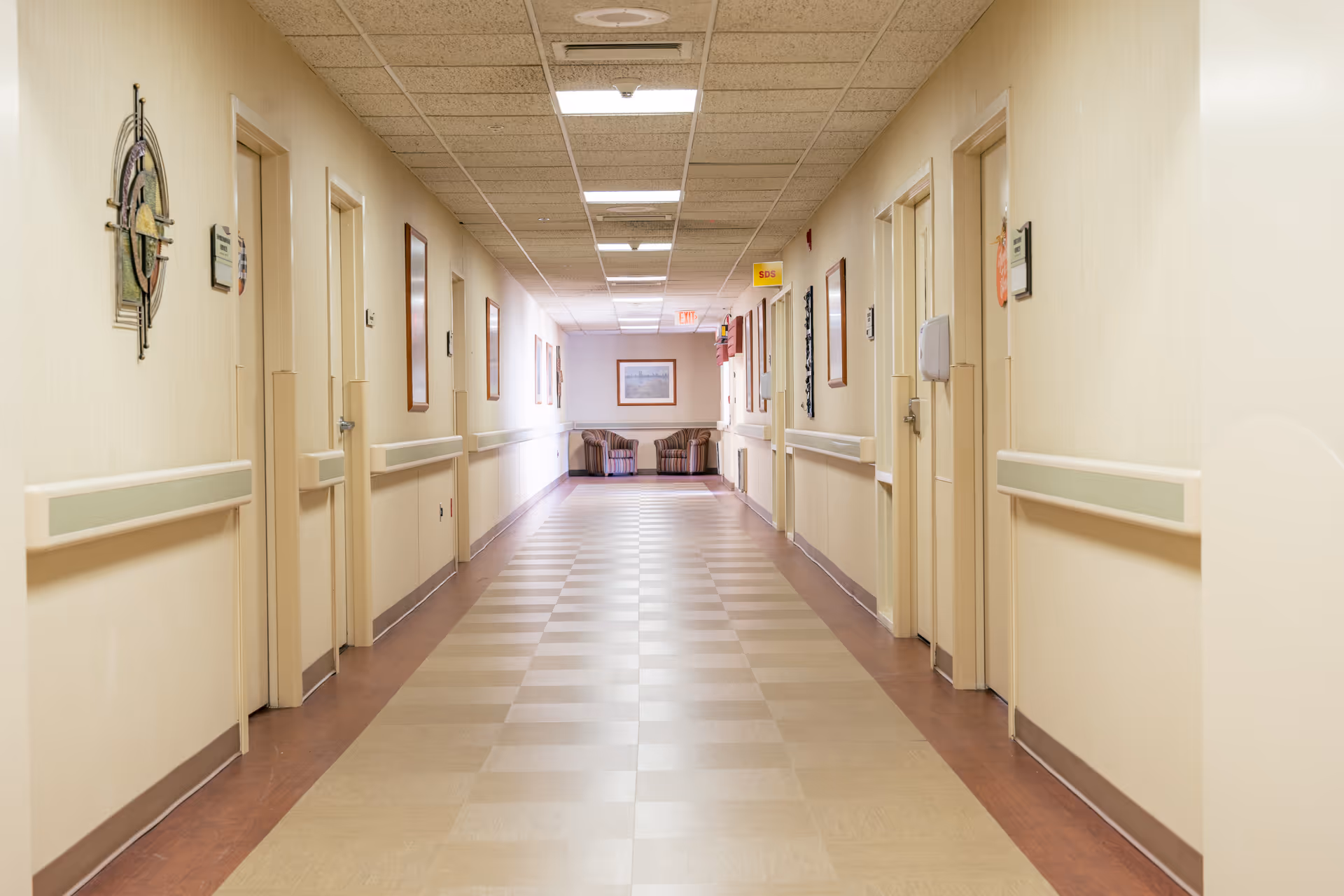 A long, well-lit hallway in a senior living facility with beige walls and checkered flooring. Several closed doors line both sides of the corridor, with handrails along the walls. At the end of the hallway, there are two striped armchairs and a framed picture on the wall.