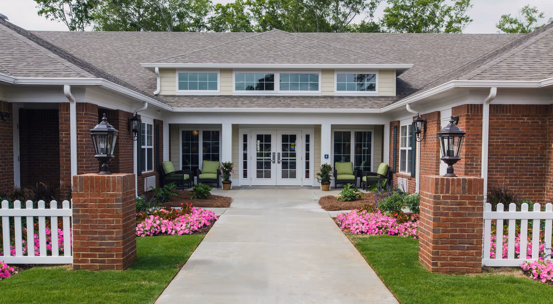 Front entrance of a senior living facility with a concrete walkway leading to double glass doors. The building has brick walls, white trim, and a gray shingled roof. There are green cushioned chairs on either side of the entrance, potted plants, and flower beds with pink flowers along the walkway. Two brick pillars with black lanterns flank the walkway, and white picket fencing is visible on both sides.