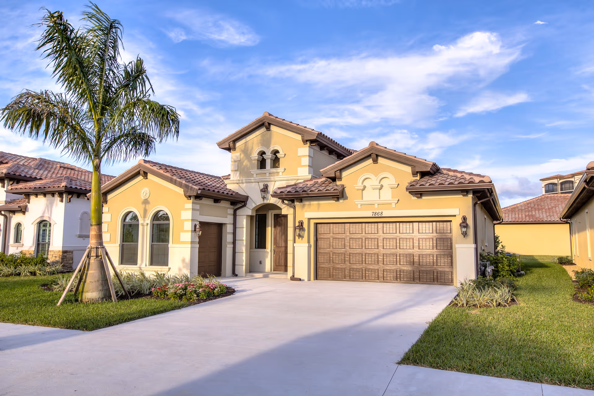 Exterior view of a single-story residential building with a tiled roof, arched windows, a double garage door, and a palm tree in the front yard under a partly cloudy blue sky.