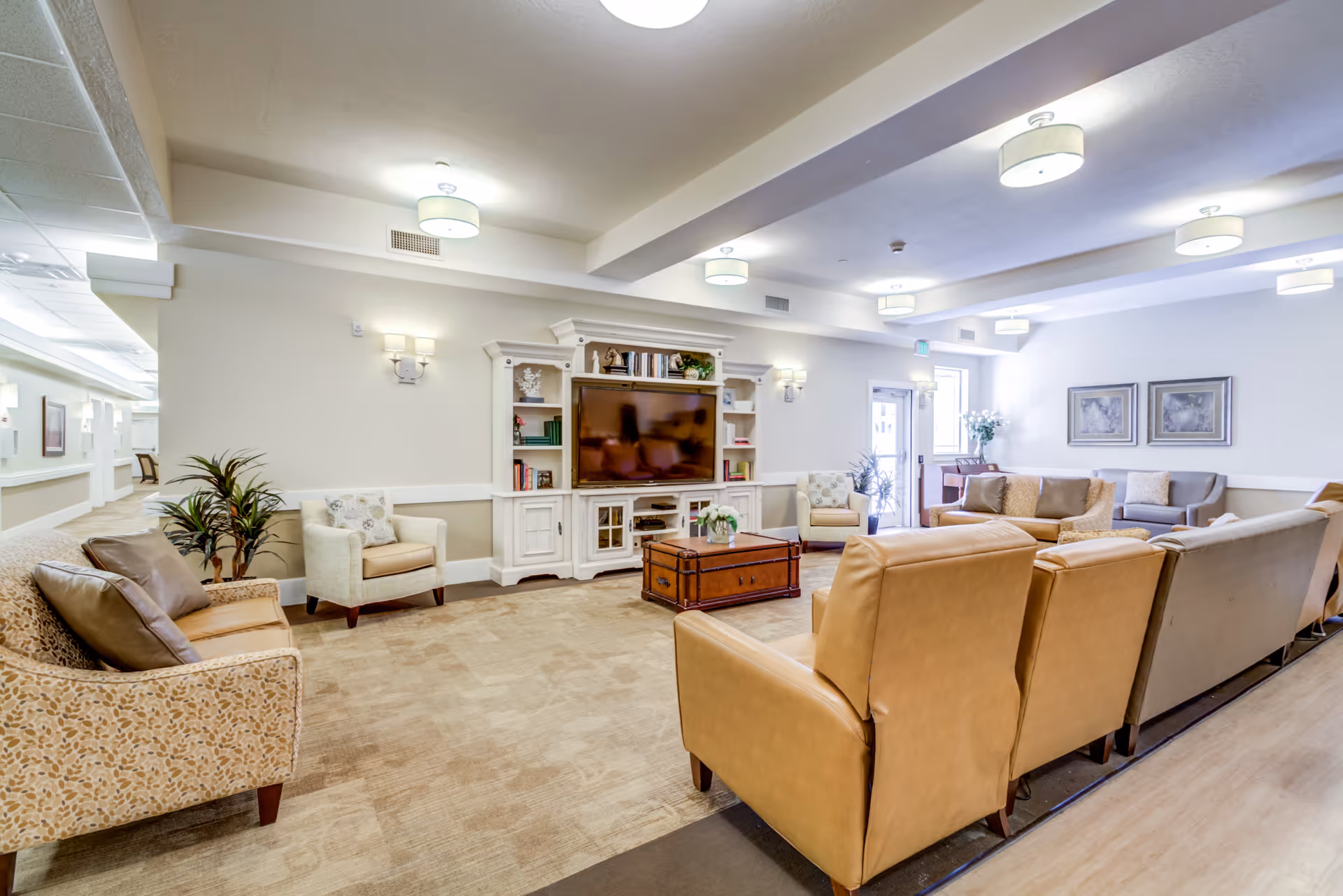 A spacious senior living facility common area with multiple comfortable seating options including beige and patterned armchairs and sofas arranged around a wooden coffee table. A large white entertainment center with a flat-screen TV and shelves filled with books and decorative items is against the wall. The room is well-lit with ceiling lights and wall sconces, and there are framed artworks and plants adding to the cozy atmosphere.