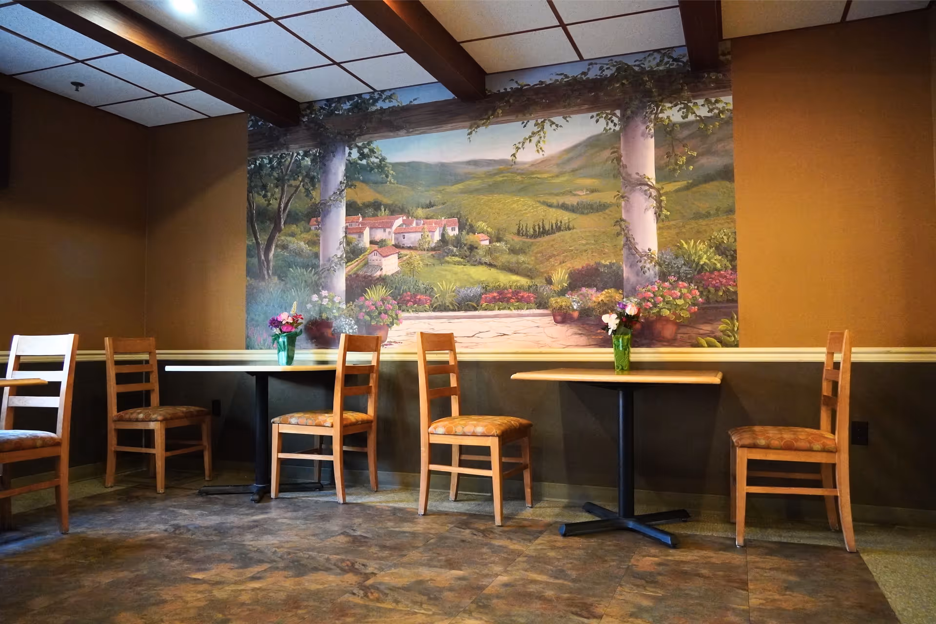A dining area with wooden chairs and tables, each table decorated with a small vase of flowers. The wall features a large mural depicting a scenic countryside landscape with rolling hills, trees, and a village in the distance.