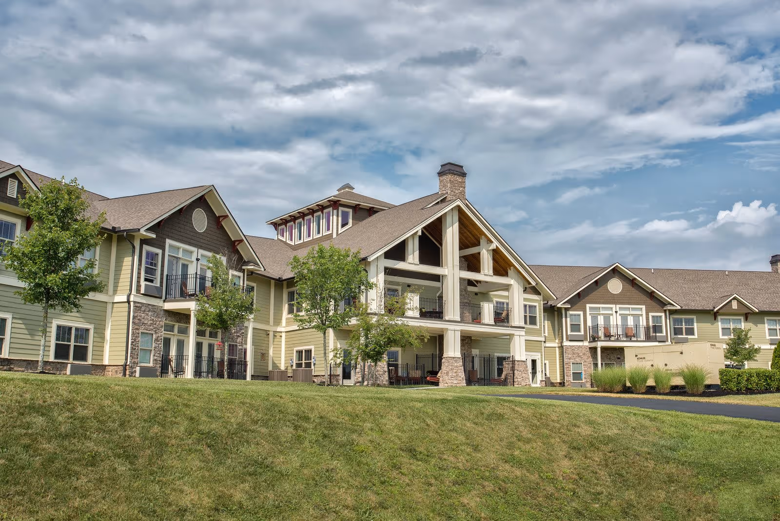 Exterior view of a large, multi-story senior living facility building with beige and brown siding, multiple balconies, and a well-maintained grassy lawn in the foreground under a partly cloudy sky.