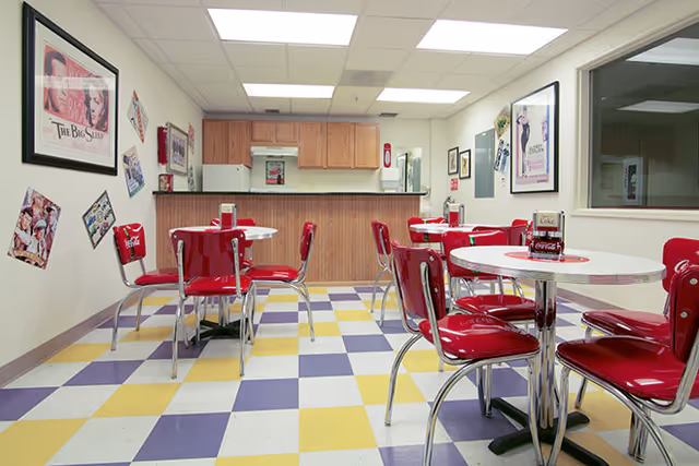 A retro-style dining area with red chairs and round white tables on a checkered yellow, white, and purple floor. The walls are decorated with vintage posters and advertisements. A wooden counter and kitchen area are visible in the background.