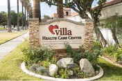 Outdoor view of the Villa Health Care Center sign surrounded by landscaping with rocks, plants, and palm trees along a sidewalk and street.