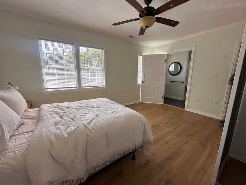 A bright bedroom with a large bed covered in white bedding, wooden floor, a ceiling fan, and two windows letting in natural light. An open door leads to a bathroom with a round mirror visible on the wall.