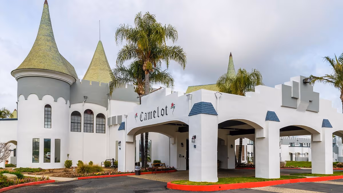 Exterior view of Camelot - A Provincial Senior Living Community featuring castle-like architecture with white walls, green conical roofs, palm trees, and an arched entrance with the name 'Camelot' displayed above.