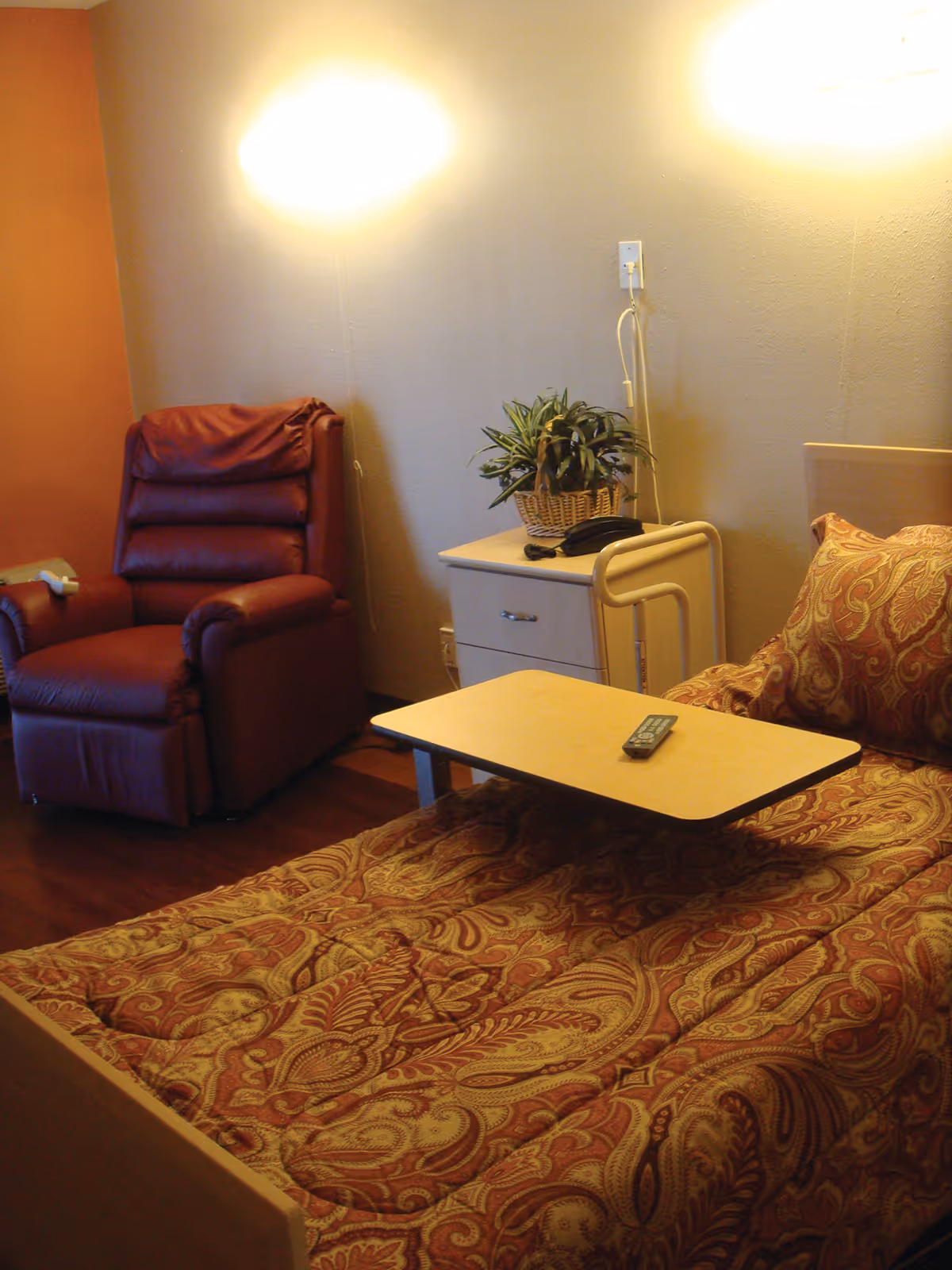 A senior living facility bedroom with a single bed covered in a patterned red and gold comforter and matching pillow. Next to the bed is a small white nightstand with a potted plant and a black telephone. A beige overbed table with a remote control on it is positioned over the bed. To the left of the nightstand is a red recliner chair. The walls are painted in neutral tones with two wall-mounted lights illuminating the room.