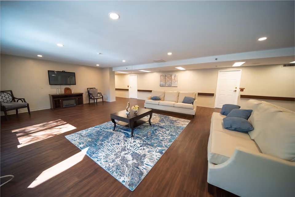 A spacious living room with two beige sofas adorned with blue cushions, a dark wooden coffee table on a blue and white patterned rug, two armchairs against the wall, a flat-screen TV mounted above a wooden cabinet, and recessed ceiling lights illuminating the room.