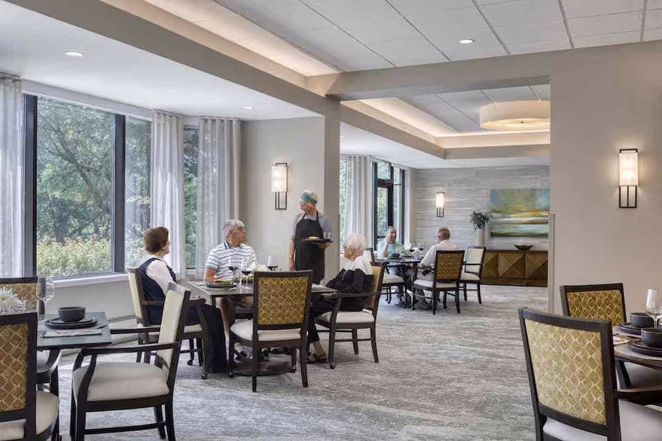 Bright dining room with seniors seated at tables and a server attending to them near large windows.