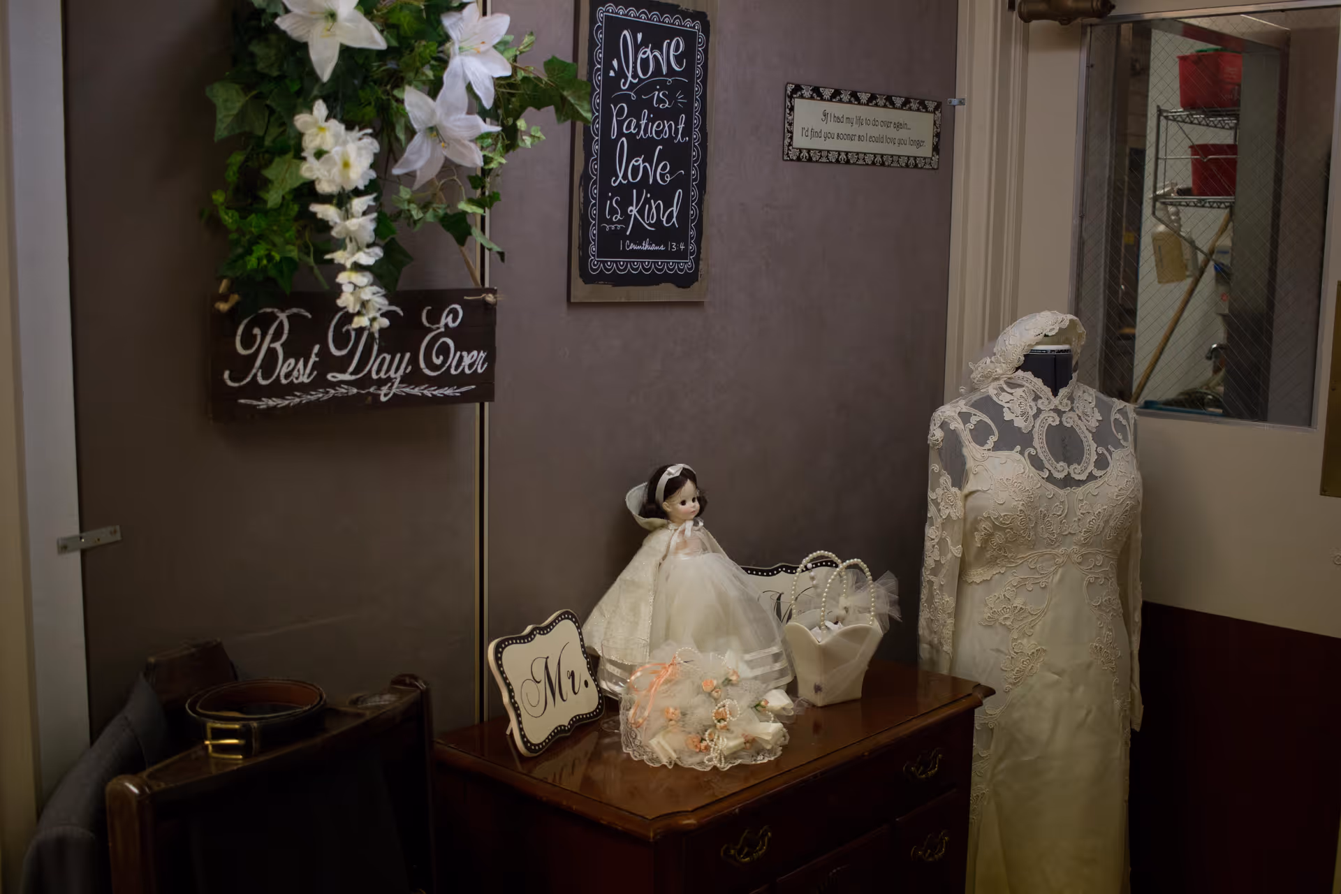 A small corner of a room decorated with a wooden chest of drawers holding a doll dressed in a white gown, a bouquet of flowers, a small white handbag, and a sign with 'Mr.' on it. To the right, there is a mannequin wearing a lace wedding dress. On the wall above, there are two framed signs, one reading 'Love is Patient, Love is Kind' and the other with a quote. A wooden box with white flowers and the words 'Best Day Ever' hangs on the wall to the left.