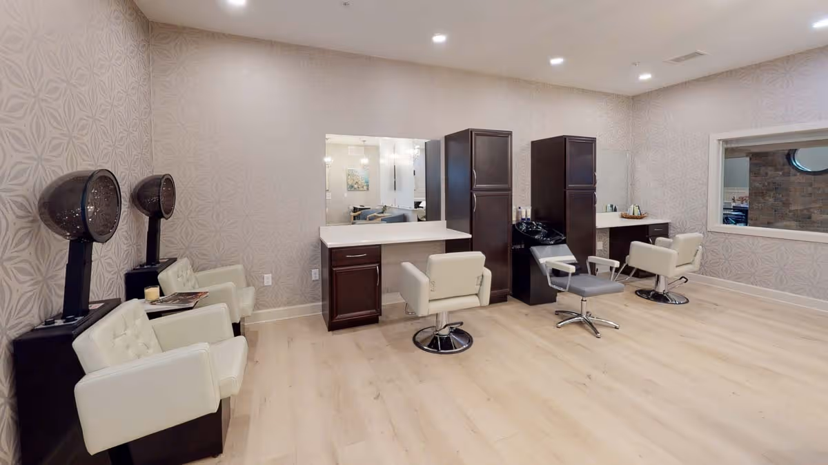 Interior view of a salon area with three styling stations featuring large mirrors, dark wood cabinets, and white swivel chairs. On the left side, there are two vintage-style hair dryers with white cushioned chairs underneath. The room has light wood flooring and patterned wallpaper, with recessed ceiling lights providing illumination.
