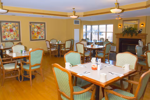 A dining room with multiple wooden tables and green cushioned chairs arranged neatly. Each table is set with white mugs, napkins, and utensils. The room has wooden flooring, yellow walls, two floral paintings, and a fireplace with a mirror and plants on the mantel. Large windows allow natural light to enter the space.