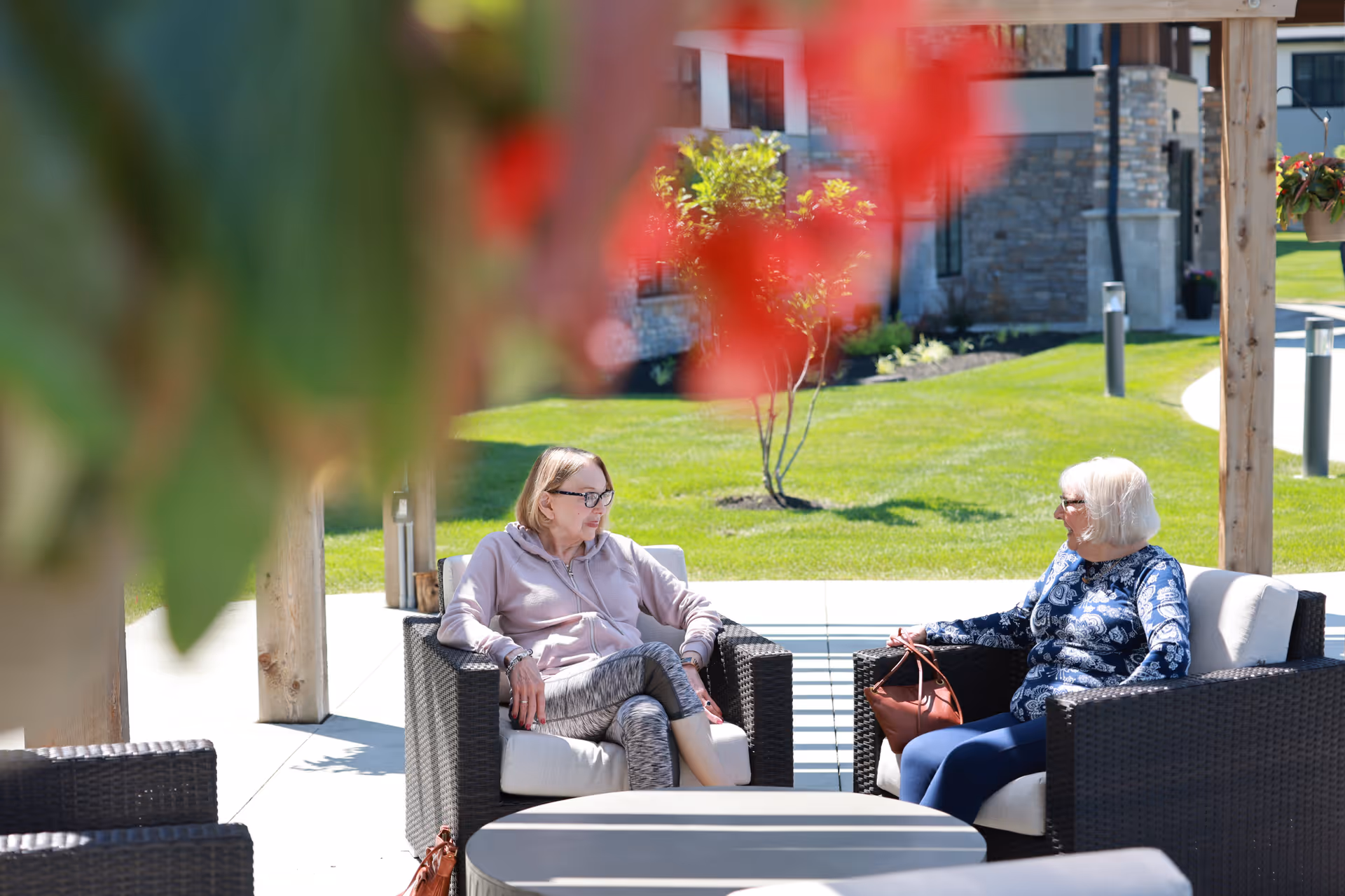 Two elderly women sitting and talking on outdoor wicker chairs with cushions under a wooden pergola, with green grass and a stone building in the background.