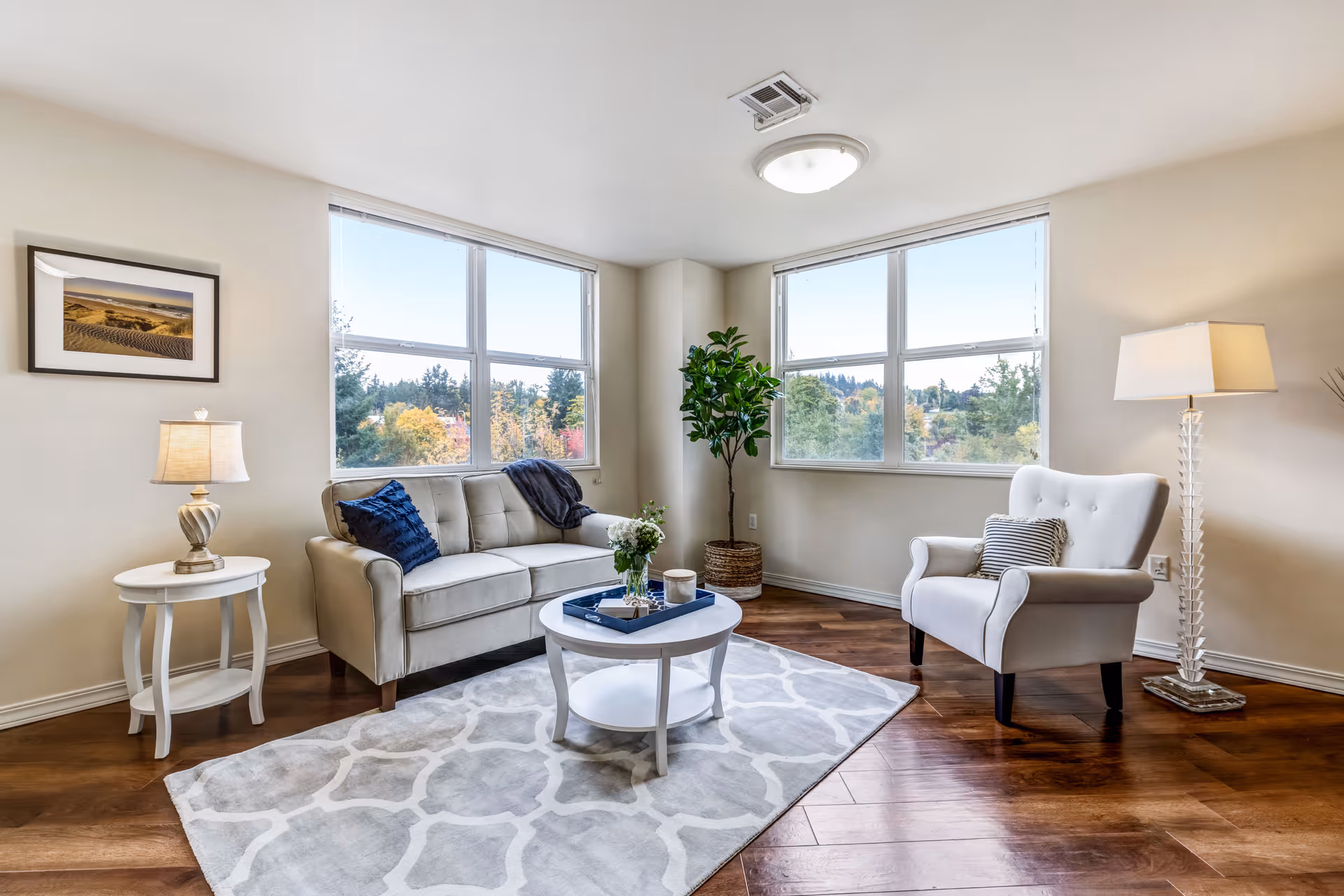 A bright and cozy living room with two large windows showing a view of trees outside. The room features a beige sofa with blue and dark gray pillows, a white armchair with a striped pillow, a white round coffee table with a blue tray and flowers, a white side table with a lamp, a tall floor lamp, a potted plant in the corner, and a framed picture on the wall. The floor is wooden with a light gray patterned rug.