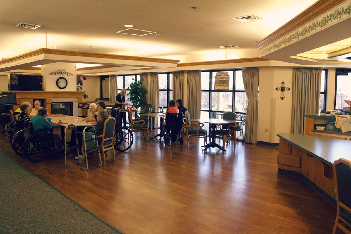A spacious common area in a senior living facility with several elderly residents sitting around tables, some in wheelchairs, engaging in activities. The room has large windows with curtains, wooden flooring, and a fireplace with a clock above it. A staff member is assisting residents in the background.