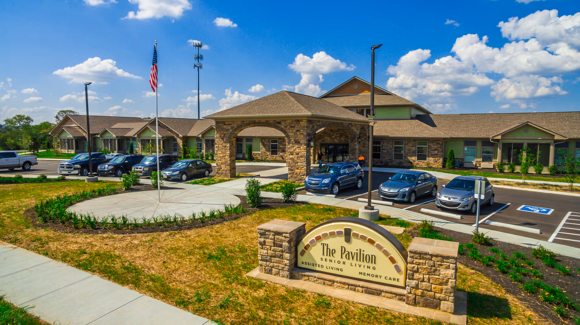 Front entrance of The Pavilion Senior Living with a stone portico, parking lot, flagpole, and the facility sign in the foreground.