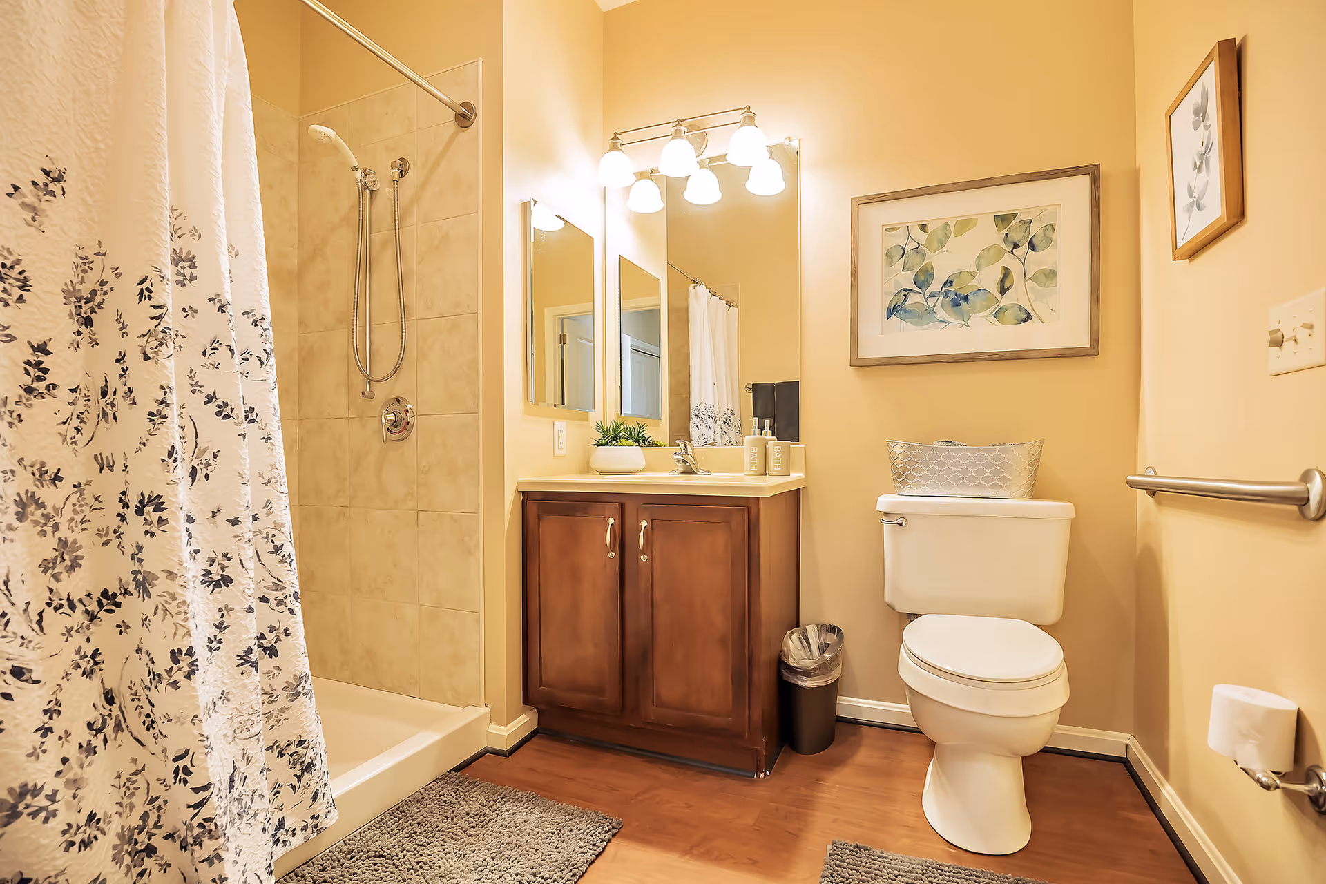A clean and well-lit bathroom featuring a shower with a floral-patterned curtain, a wooden vanity with a sink and mirror above it, a white toilet with a decorative basket on top, framed botanical artwork on the walls, and a trash can beside the vanity. The floor has wooden flooring with gray bath mats.