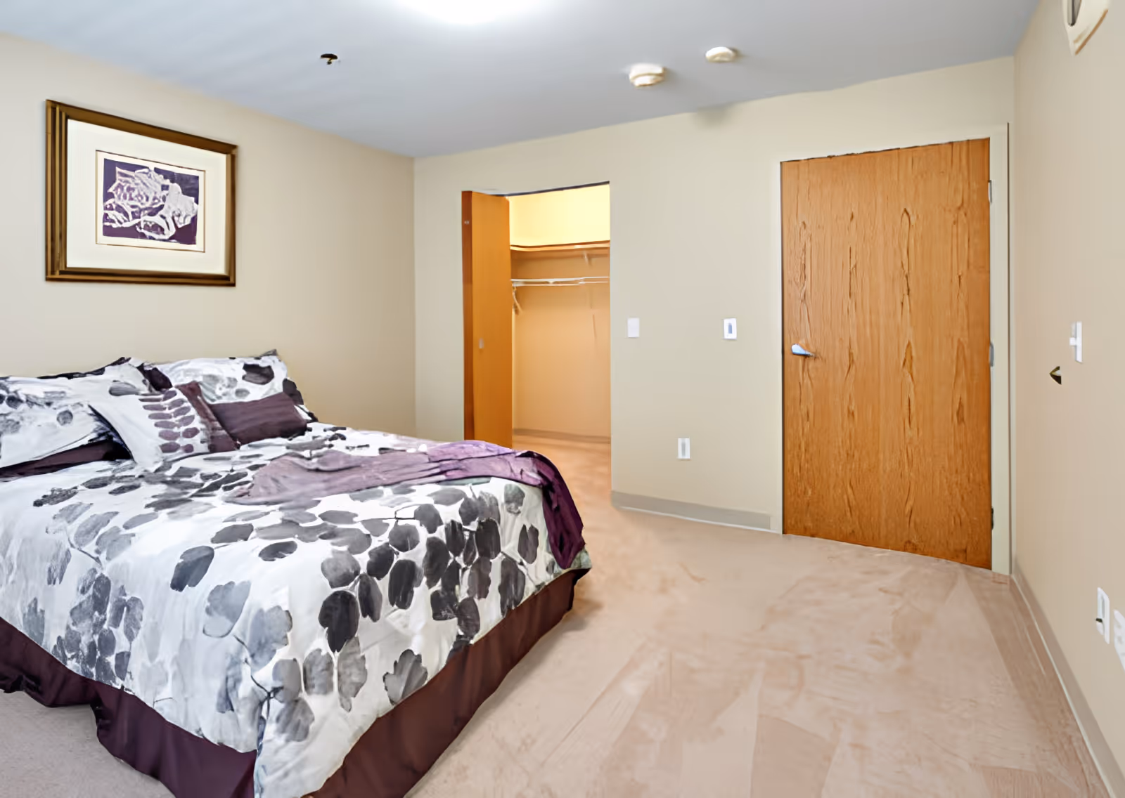 A furnished bedroom with a patterned bed, open walk-in closet, and a closed wooden door.