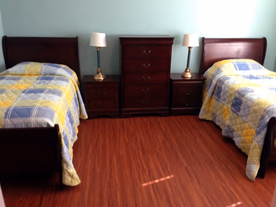 A bedroom with two twin beds, each with a yellow, blue, and gray patchwork quilt. Between the beds are two nightstands with lamps and a tall chest of drawers in the center. The floor is wooden and the walls are light blue.