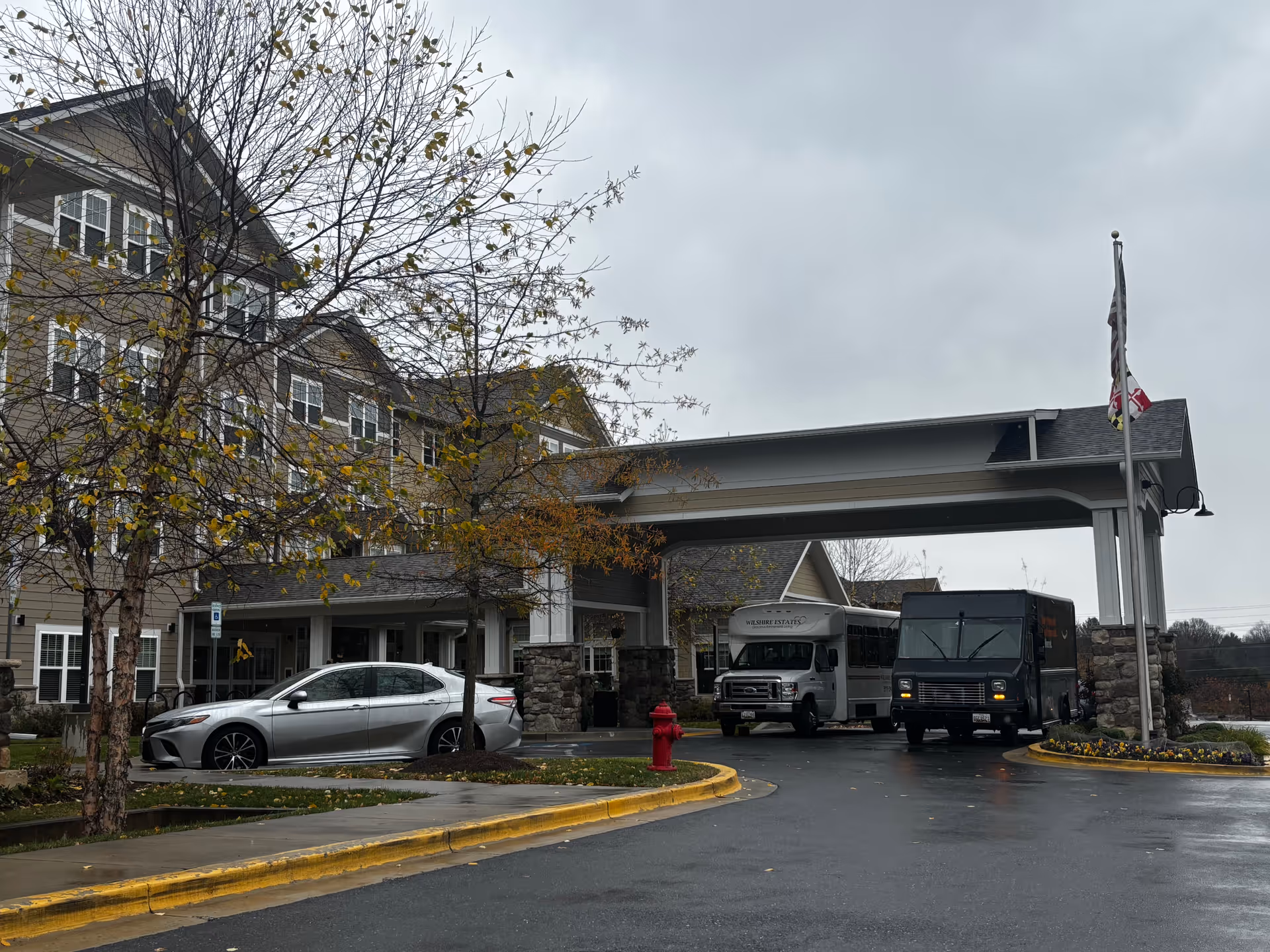 Covered porte-cochère entrance of a multi-story senior living building with parked cars, a shuttle bus, and a flag on a cloudy day.