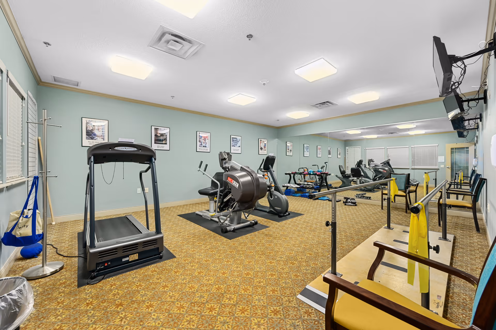 A fitness room in a senior living facility with exercise equipment including a treadmill, stationary bikes, and parallel bars for physical therapy. The room has light blue walls, patterned carpet, several framed pictures on the walls, and a large mirror covering one wall. There are chairs along the wall and a TV mounted in the corner.