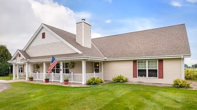 Exterior view of a single-story residential building with beige siding, a brown shingled roof, and a front porch with white railings. There is an American flag displayed on the porch, several potted plants, and well-maintained green grass surrounding the building under a partly cloudy sky.