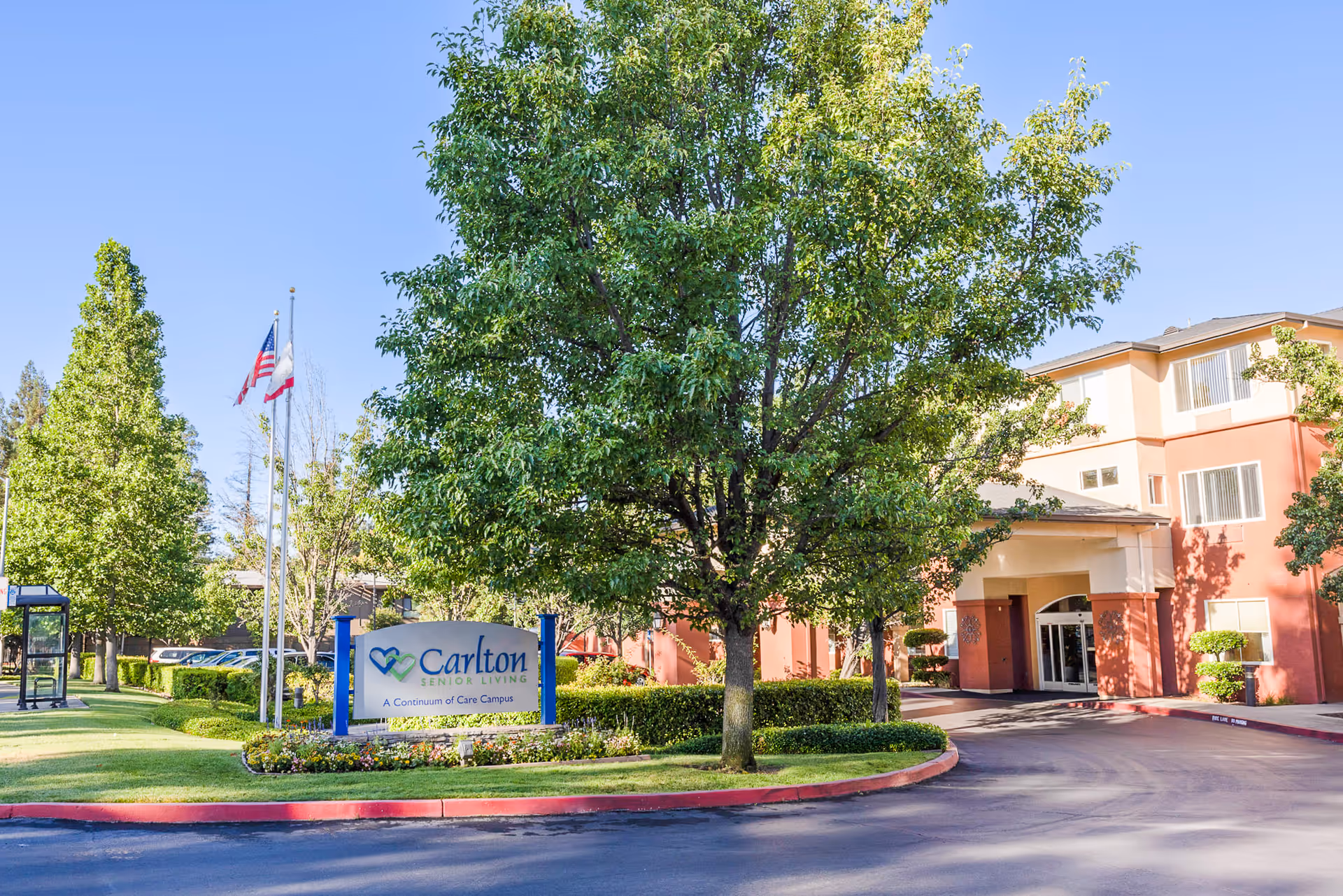 Exterior view of Carlton Senior Living Sacramento facility with a large tree, manicured bushes, and a sign that reads 'Carlton Senior Living A Continuum of Care Campus'. The building is a multi-story structure with a covered entrance and several windows. Two flagpoles with the American and California state flags are visible in the background.