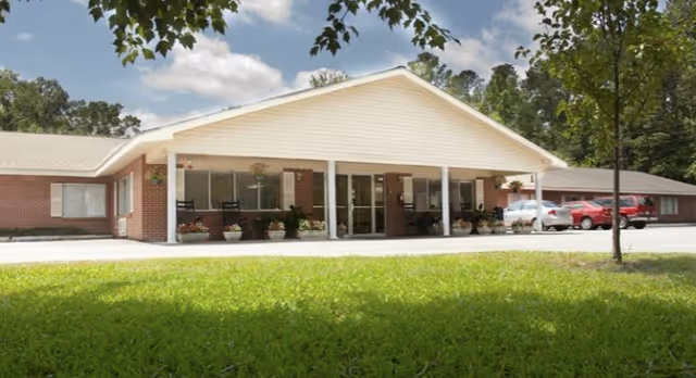 Front view of a single-story brick senior living facility with a covered entrance, potted plants, parked cars, and a grassy lawn in front.
