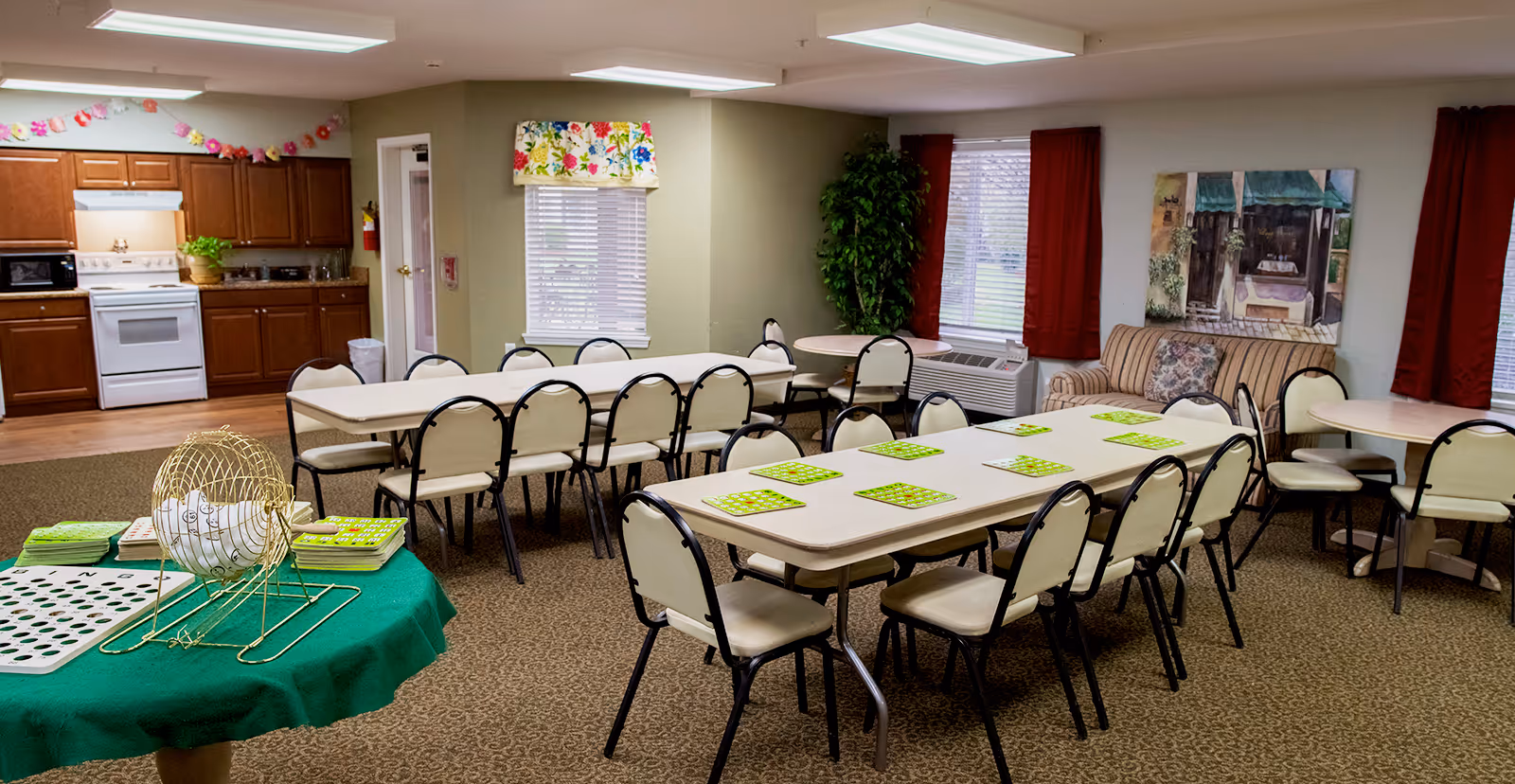 A senior living community common room with rows of tables and chairs set up for a bingo game, a bingo cage on a side table, and a kitchenette in the background.