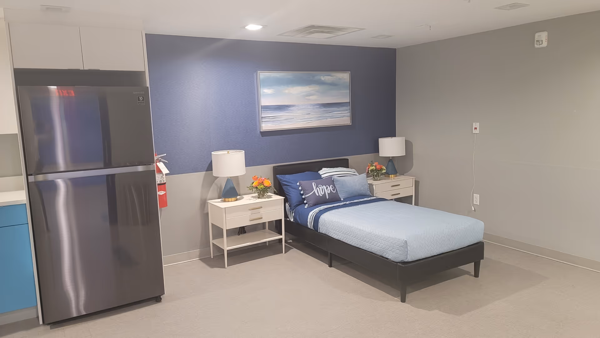 A simple room with a single bed dressed in blue bedding and a pillow with the word 'hope'. There are two white nightstands on either side of the bed, each with a lamp and a small flower arrangement. Behind the bed is a blue accent wall with a framed picture of a seascape. To the left of the bed is a stainless steel refrigerator and part of a kitchen counter with blue cabinets. The walls are painted light gray and the floor is carpeted.