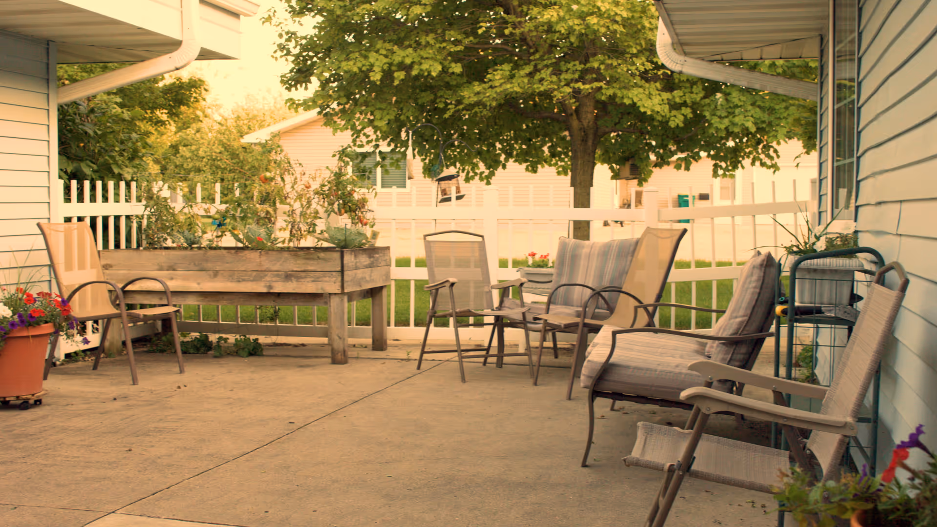 Outdoor patio area with several cushioned chairs arranged around a concrete floor, a raised wooden planter box with plants, potted flowers, and a white picket fence with trees and houses in the background.