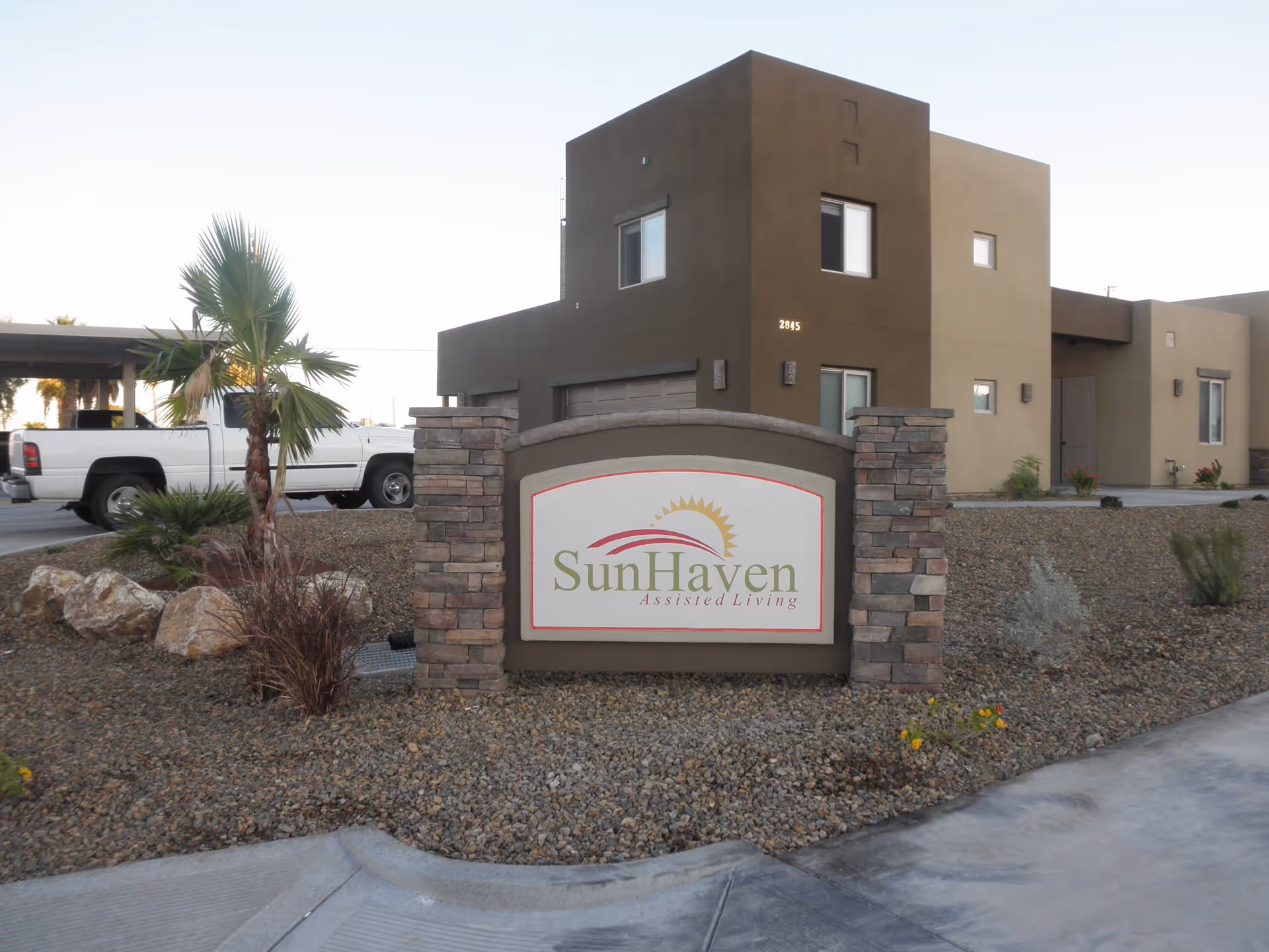 Exterior view of a modern building with a stone and stucco sign in front that reads 'SunHaven Assisted Living'. The building is surrounded by desert landscaping with rocks, small plants, and a palm tree. A white pickup truck is parked to the left side of the building.