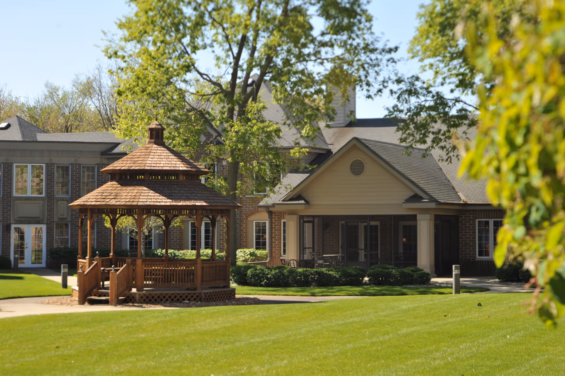 A wooden gazebo with a shingled roof stands on a well-maintained lawn in front of a senior living facility building. The building has a brick and beige exterior with multiple windows and a covered patio area with outdoor seating. Trees with green leaves surround the area under a clear blue sky.