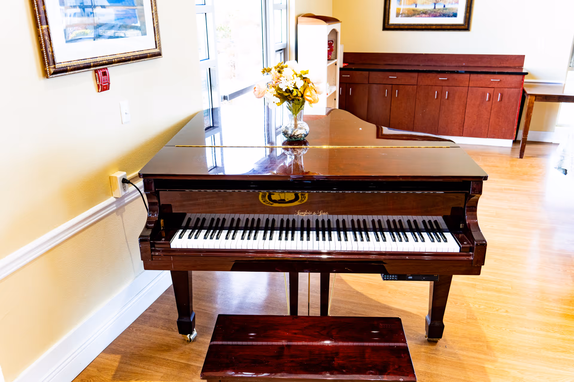 A polished wooden grand piano with a matching bench in a well-lit room with wooden flooring. A vase with yellow and white flowers is placed on top of the piano. The room has light-colored walls, a framed picture, and wooden cabinets in the background.