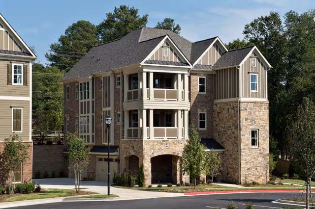 Front view of a three-story stone-and-siding senior living building with covered balconies, garage openings, and landscaped grounds.