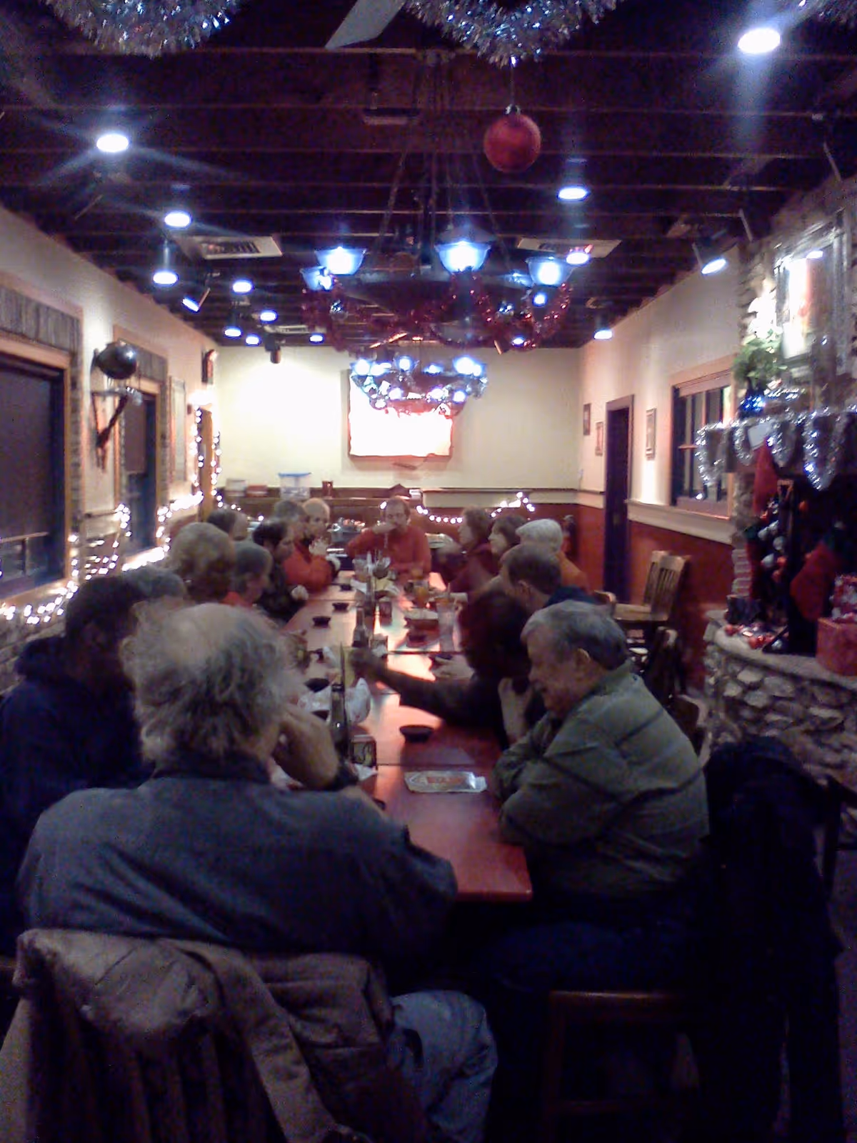A group of elderly people sitting around a long dining table in a warmly lit room decorated with Christmas ornaments and string lights. The room has wooden beams on the ceiling and a stone fireplace adorned with holiday decorations on the right side.